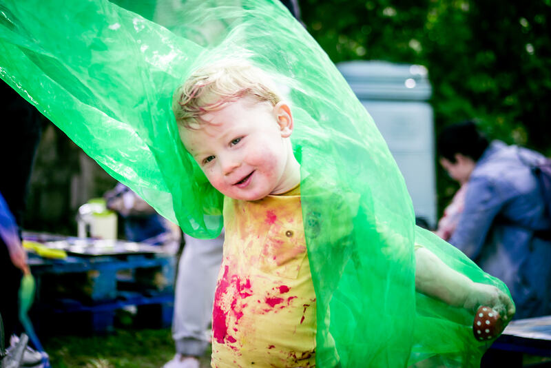 A smiling toddler emerges from under a piece of hanging green organza, arms outstretched. Smatterings of red paint cover their hair and clothes. Clutched in their hand - just visible through the organza - is a tiny spotty red teacup