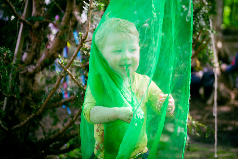 A smiling child has wrapped himself in a piece of green organza which hangs from a tree.