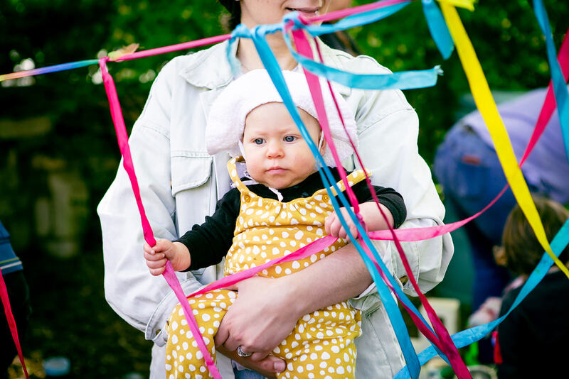 A baby wearing a floppy sun hat, held in the arms of their grown-up. They clutch inquisitively at the pink, blue and yellow ribbons hanging in front of them.