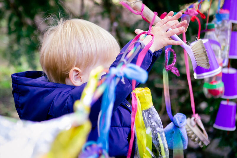 A toddler reaches up to grab a washing up brush that hangs from a ribbon, alongside other colourful dangling objects, plastic bottles and paper cups.
