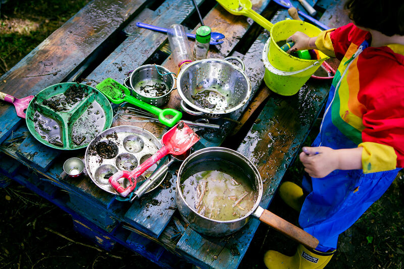 Birds eye view of a child in a rainbow rainsuit playing with a mud kitchen - plastic spades, saucepans, buckets and plates full of water sit atop a blue wooden pallet.