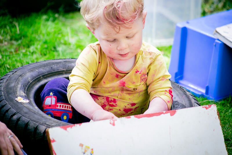 A toddler sits inside a tyre, wearing a yellow top covered in splatters of red paint.