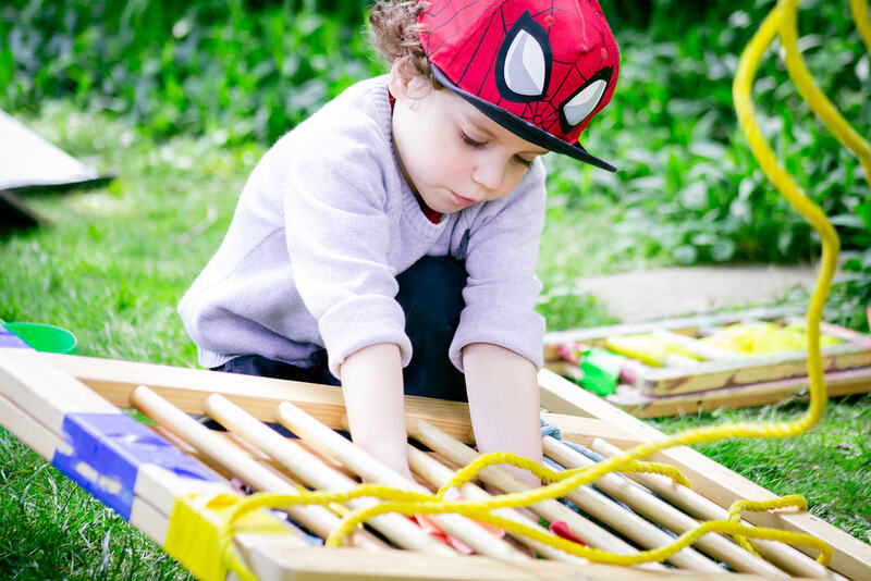 A child wearing a Spider Man hat kneels over an old bed frame, weaving a bright yellow rope through the slats.