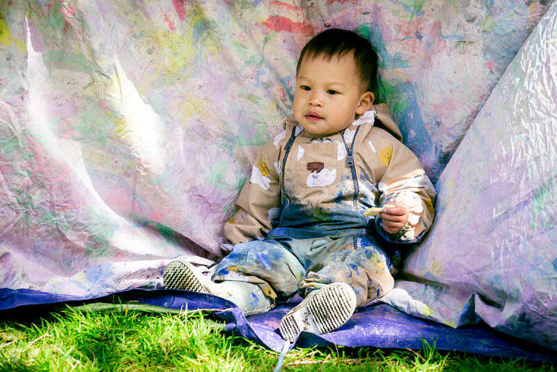 A toddler in a rainsuit covered in paint sits against a paint stained sheet.