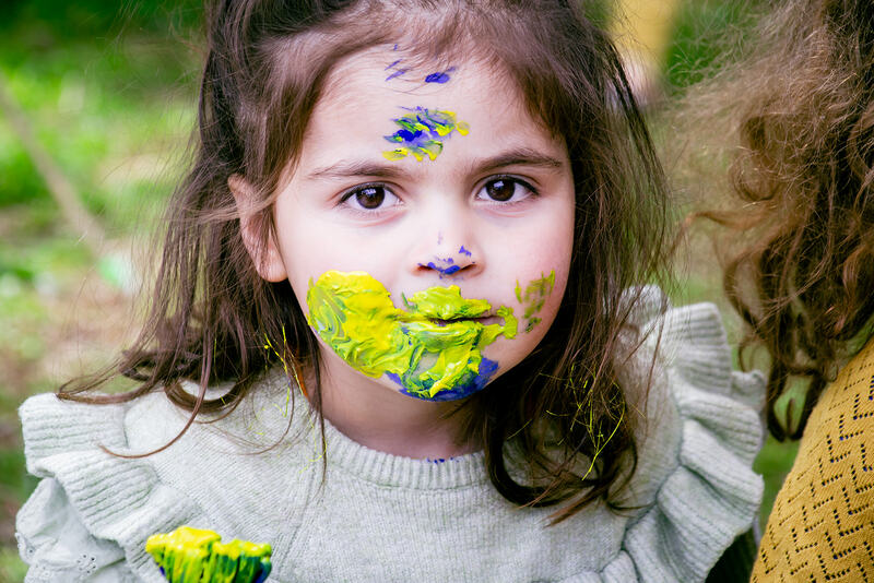 A child looks at the camera, her face covered in yellow and blue paint.