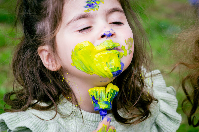 A child presses a paintbrush to her neck. Her face is coated in yellow and blue paint, her eyes are closed.