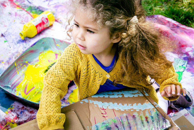 A child leans on a cardboard box, a tray of yellow paint behind her.