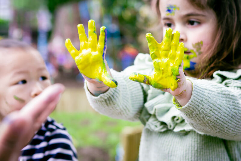 A child holds up their hands, which are slathered in yellow paint.
