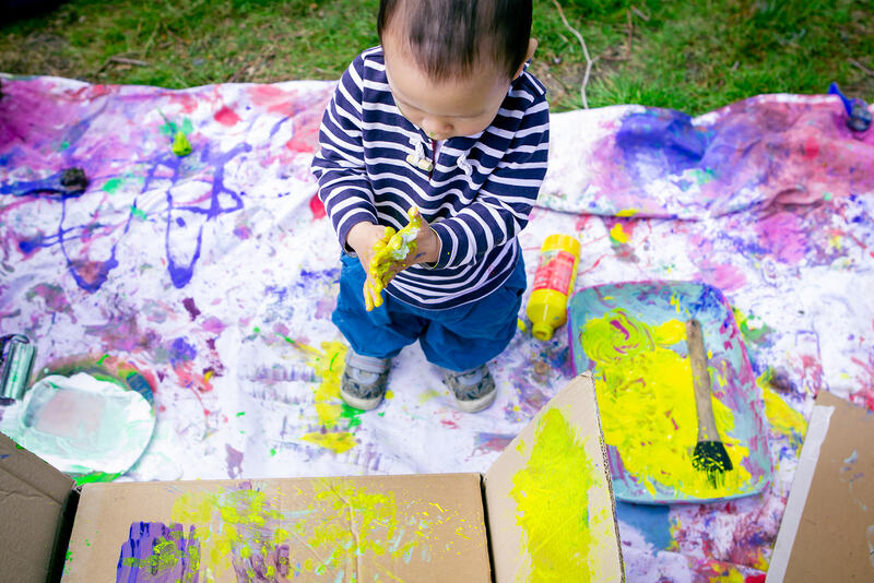 Birds eye view of a toddler standing on a paint stained sheet next to a tray of paint, rubbing yellow paint into their hands.