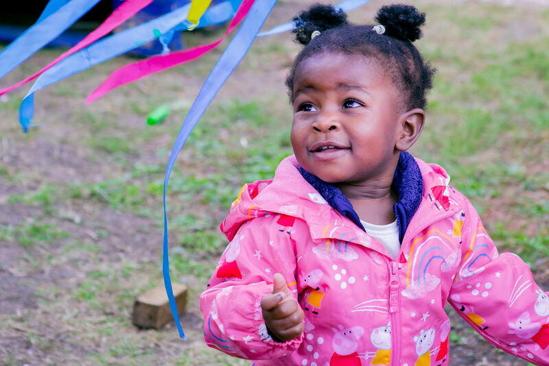 A toddler in a bright pink rain suit looks up, smiling. Blue and pink ribbons float by her head. 