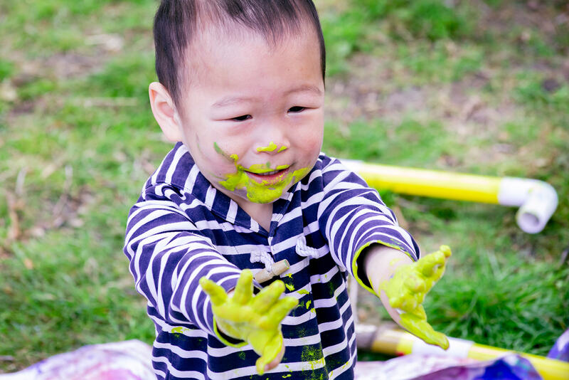 A smiling toddler with a face smeared in yellow paint holds out his hands which are also covered in paint. 
