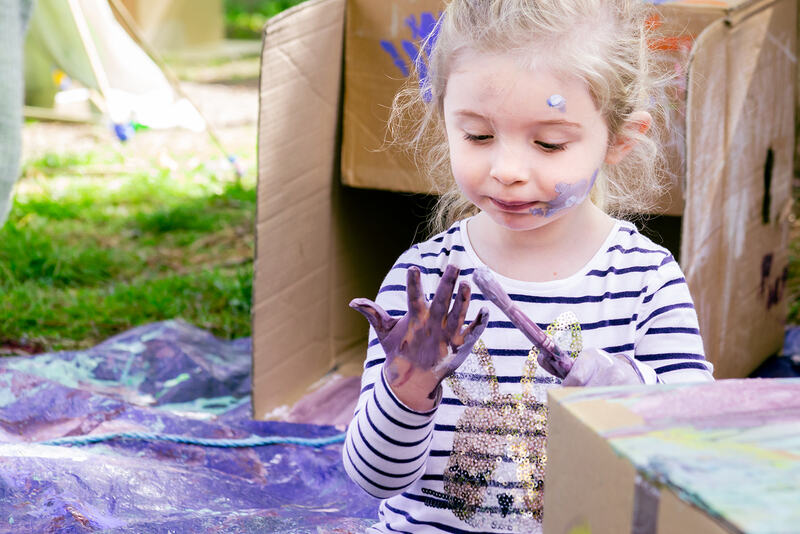 A child sits in front of a cardboard box, lilac paint smeared on her face. She clutches a paintbrush and holds her other lilac painted hand up in front of her.