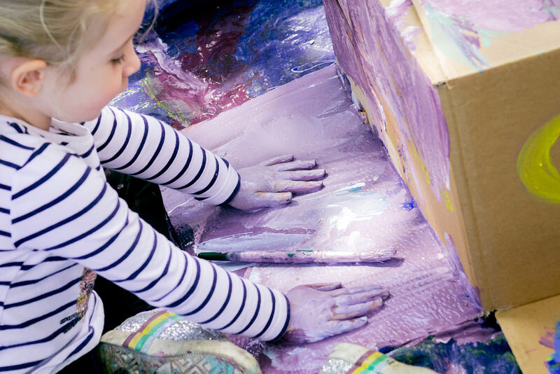 A child rubs lilac paint onto a cardboard box with their hands.
