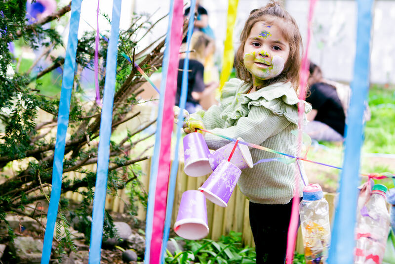 A child stands amongst a curtain of blue and pink ribbons. Her smiling face is covered in white, green and blue paint and she holds onto a string of dangling bottles and purple paper cups. 