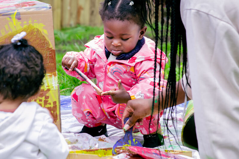 A toddler in a pink rainsuit crouches on a paint stained sheet, tentatively touching the tip of her finger with a painty paintbrush.