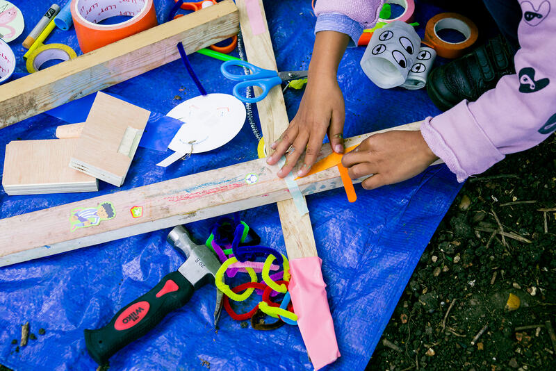 Craft materials lie on a blue tarpaulin. A hammer, tape, scissors, pipe cleaners, offcuts of wood, googly eyes. A child’s hands press tape onto a piece of wood.
