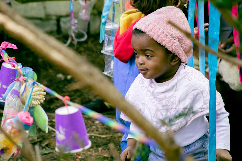 A toddler in a pink beanie hat hurries through the garden, amongst branches strung with rainbow ribbons, paper cups, plastic bottles and brushes.