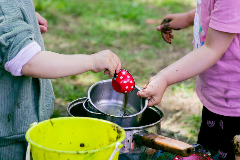 Close-up of one child pouring a teacup of water into a saucepan held by another child.