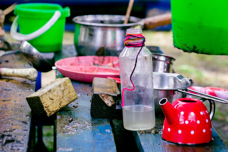 Close up of mud kitchen utensils on a muddy blue wooden pallet - a red teapot, plastic bottle, offcuts of wood, two green buckets, saucepans and a pink plate.