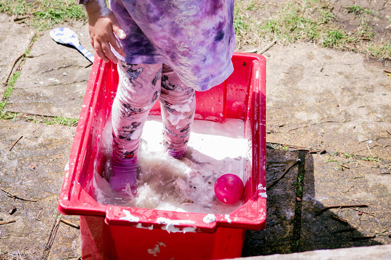 A child’s legs, wearing leggings and socks, stand in a red plastic box full of white foamy water.
