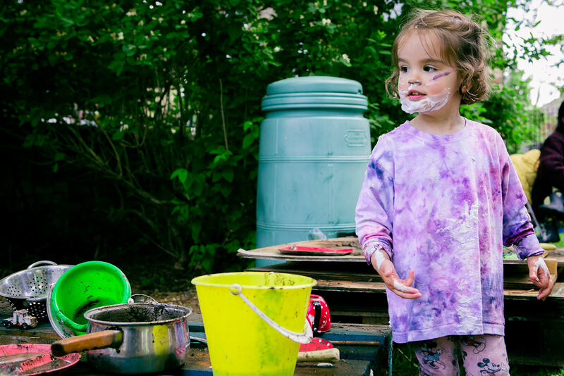 A child in a lilac tie-dye top stands in front of a pile of wooden boards and a compost bin. Her face and hands are streaked with shaving foam and lilac paint. A yellow bucket, saucepan and red teapot are in the foreground.