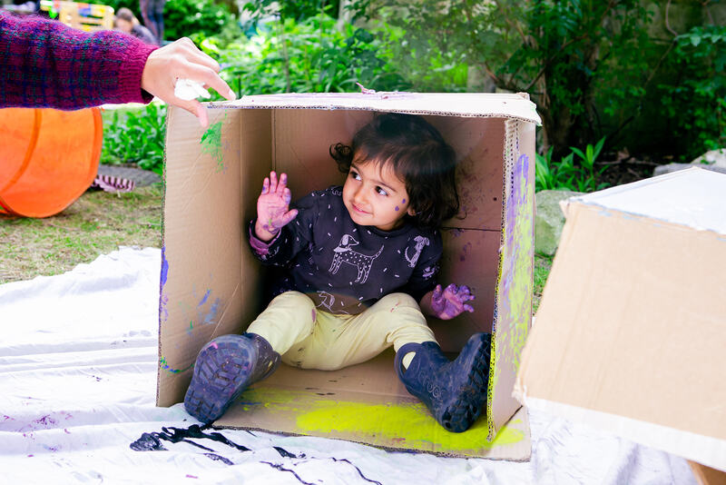 An adult hand lifts up the flap of a cardboard box to reveal a smiling child with hands covered in paint.