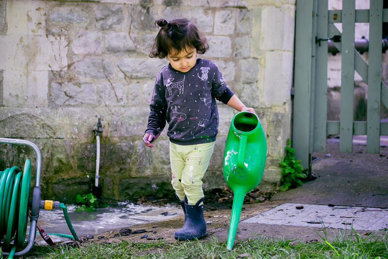 A child wearing wellies stands holding a large green watering can. Next to her is a garden hose and behind her on the wall is an outdoor tap.