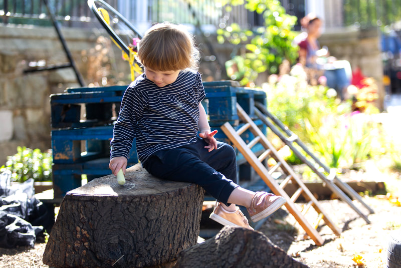 A child sits on a tree stump, drawing on it with chalk. In the background is a pile of wooden pallets with ladders propped against them.