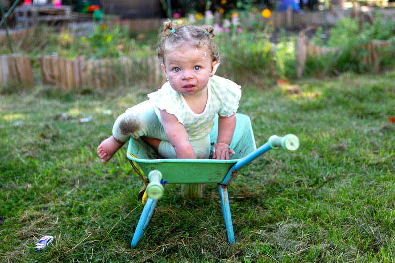 A barefoot toddler with muddy knees clambers into a small green wheelbarrow. She has smatterings of earth and foam on her face and looks into the camera.