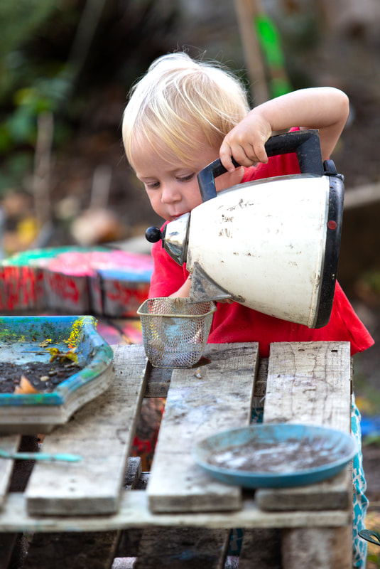 A toddler plays with a kettle, pretending to pour it into a container.
