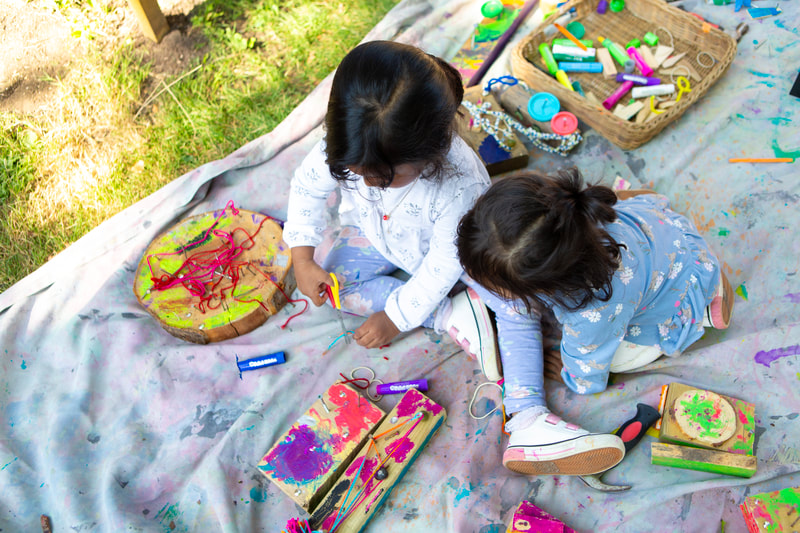 Two children sit crafting on a large sheet, surrounded by pieces of wood, paintsticks, wool and tools