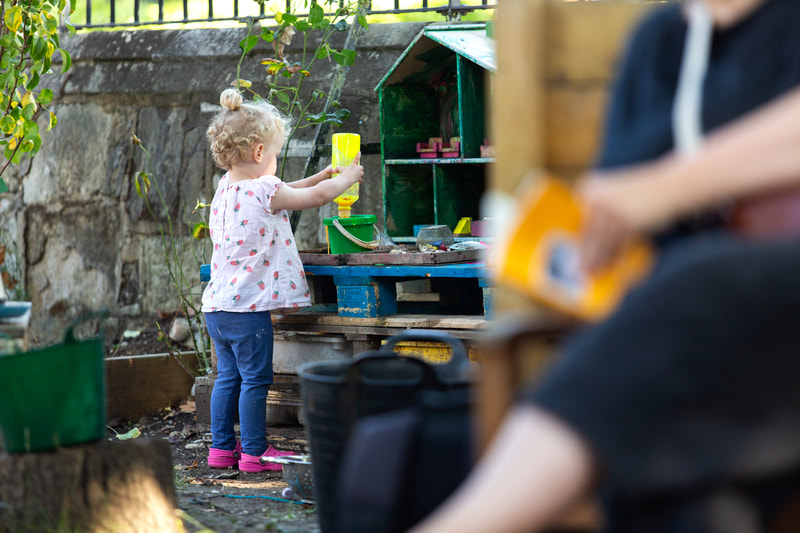 A child stands by a pile of pallets with a dollhouse on top, squeezing yellow paint into a bucket. In the foreground we can just see an adult sitting reading a book.