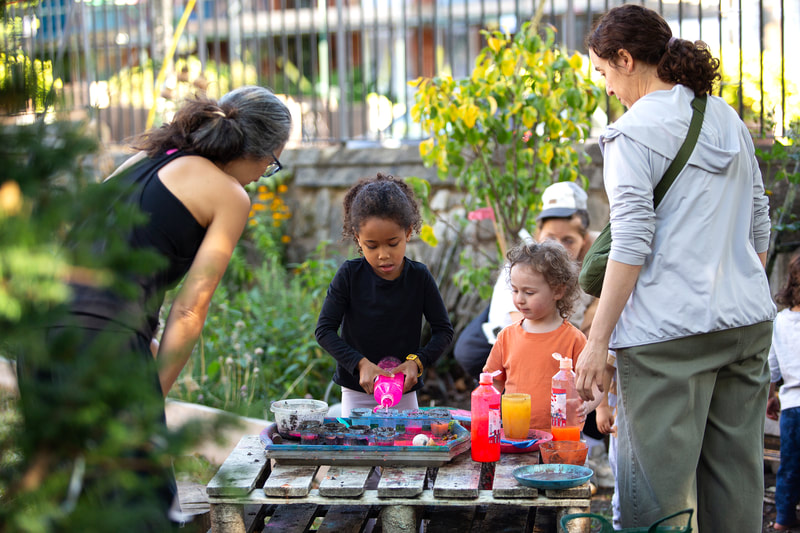 Children and adults gather round a wooden pallet table as a child squeezes pink water into kitchen pots.