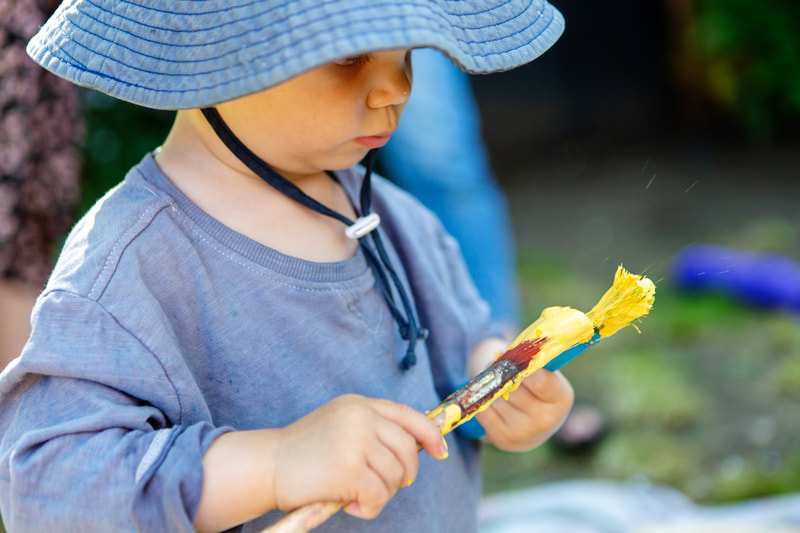 A toddler holds two paintbrushes covered in yellow paint.