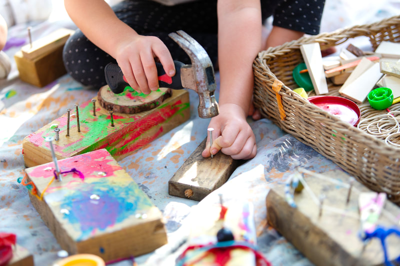 A child's hands hammer a nail into a piece of wood, surrounded by more pieces of wood and a basket of craft materials.