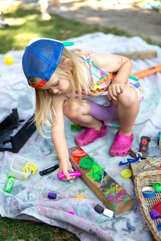 A child crouches on a sheet covered in tools and craft materials, colouring in a piece of wood with paint sticks.